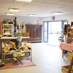 Winkelvloer van kringloopwinkel met meubels, kasten, een eettafel, kleding aan hangers en diverse huishoudelijke spullen.