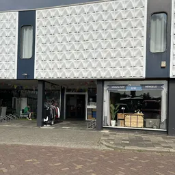 Gevel van kringloopwinkel met donkerblauwe puien, etalages met tweedehands spullen en plantenpot zichtbaar