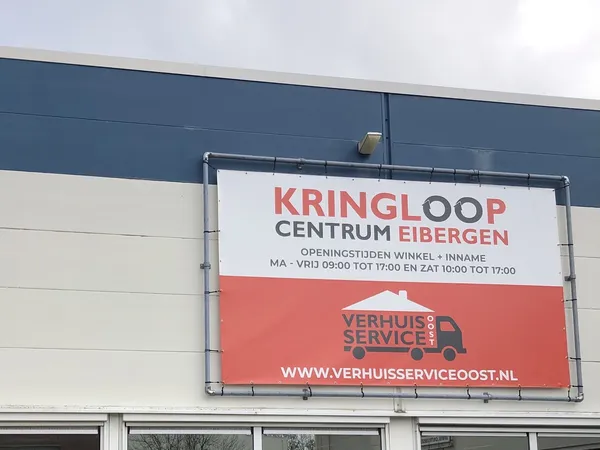 Gevel van kringloopwinkel met groot wit uithangbord en logo, onderaan rood servicelogo zichtbaar