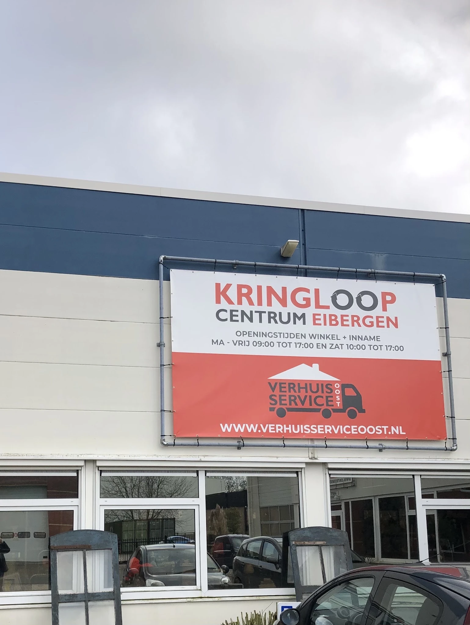 Gevel van kringloopwinkel met groot wit uithangbord en logo, onderaan rood servicelogo zichtbaar
