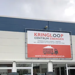 Gevel van kringloopwinkel met groot wit uithangbord en logo, onderaan rood servicelogo zichtbaar