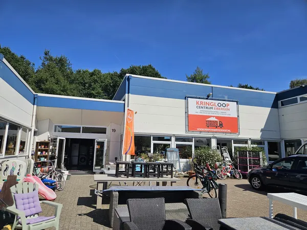 Voorkant van kringloopwinkel met blauwe en witte gevel, oranje banner, terras met stoelen en fietsen