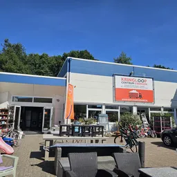 Voorkant van kringloopwinkel met blauwe en witte gevel, oranje banner, terras met stoelen en fietsen