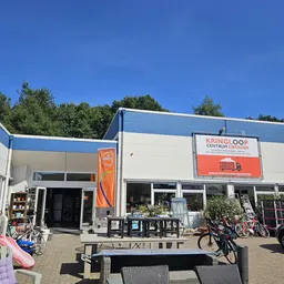 Voorkant van de kringloopwinkel met blauwe en witte gevel, grote ramen, oranje banner en winkelnaam op bord, fietsen ervoor