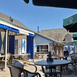 Terras met tafels, stoelen en blauwe parasols bij een bakstenen gebouw met groene luifels