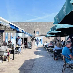 Terras met blauwe parasols waar bezoekers zitten te eten en drinken in de zon