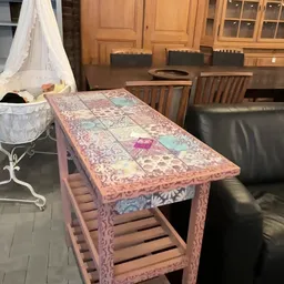 Roze beschilderde houten tafel met kleurig tegelwerk op het blad, in een interieur met houten stoelen op de achtergrond