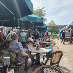 Terras met bezoekers onder blauwe parasol, tafels en stoelen op houten platform