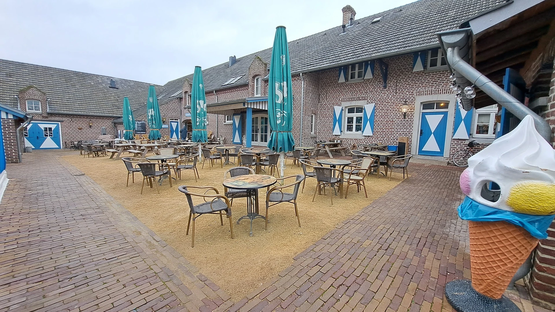Terras met tafels, stoelen en blauwe parasols tegen een bakstenen gebouw
