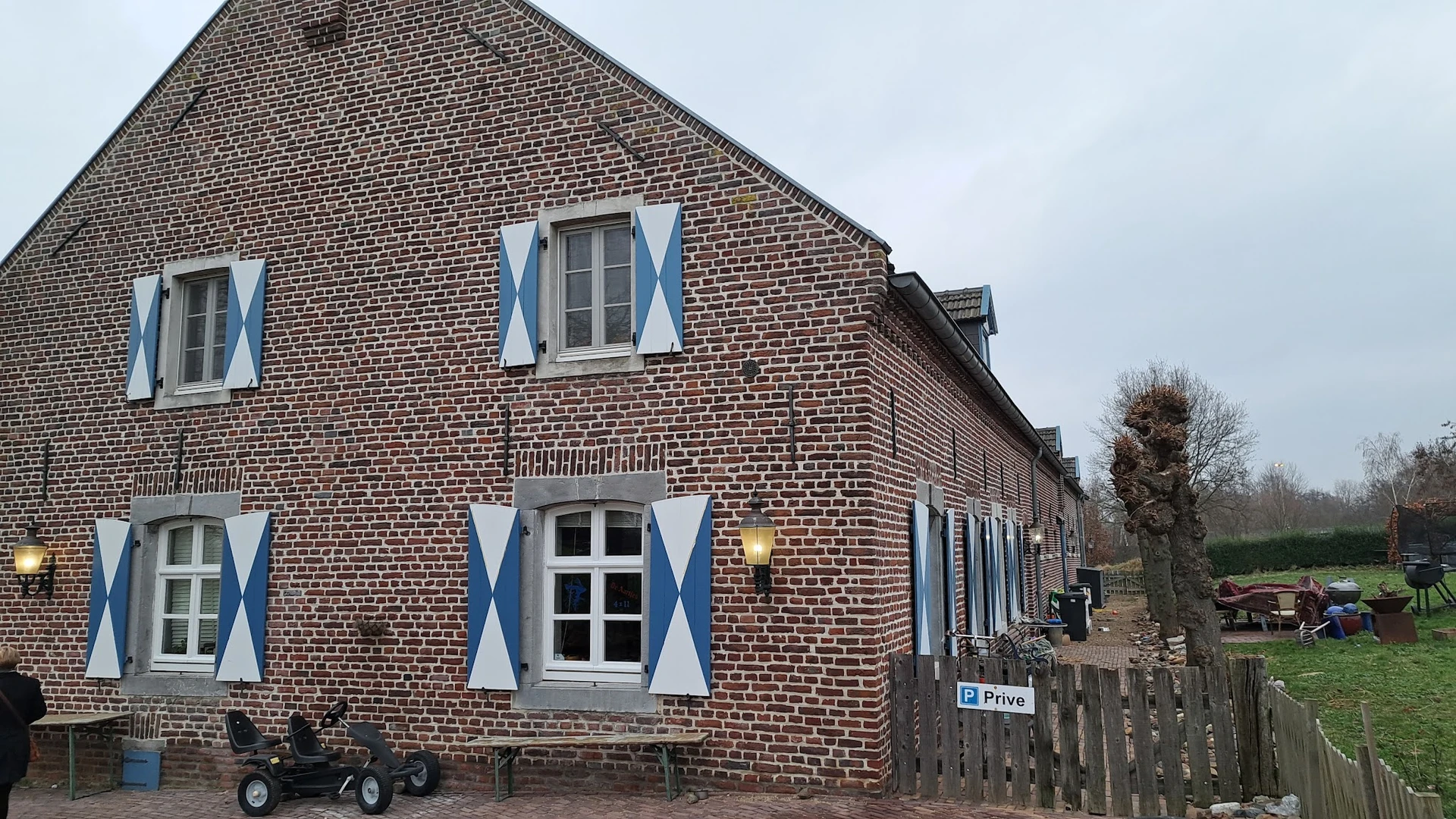 Voorgevel van bakstenen woonhuis met blauwe luiken en witte raamomlijstingen, wit deuren beneden, houten omheining vooraan