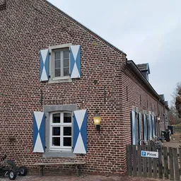 Voorgevel van bakstenen woonhuis met blauwe luiken en witte raamomlijstingen, wit deuren beneden, houten omheining vooraan