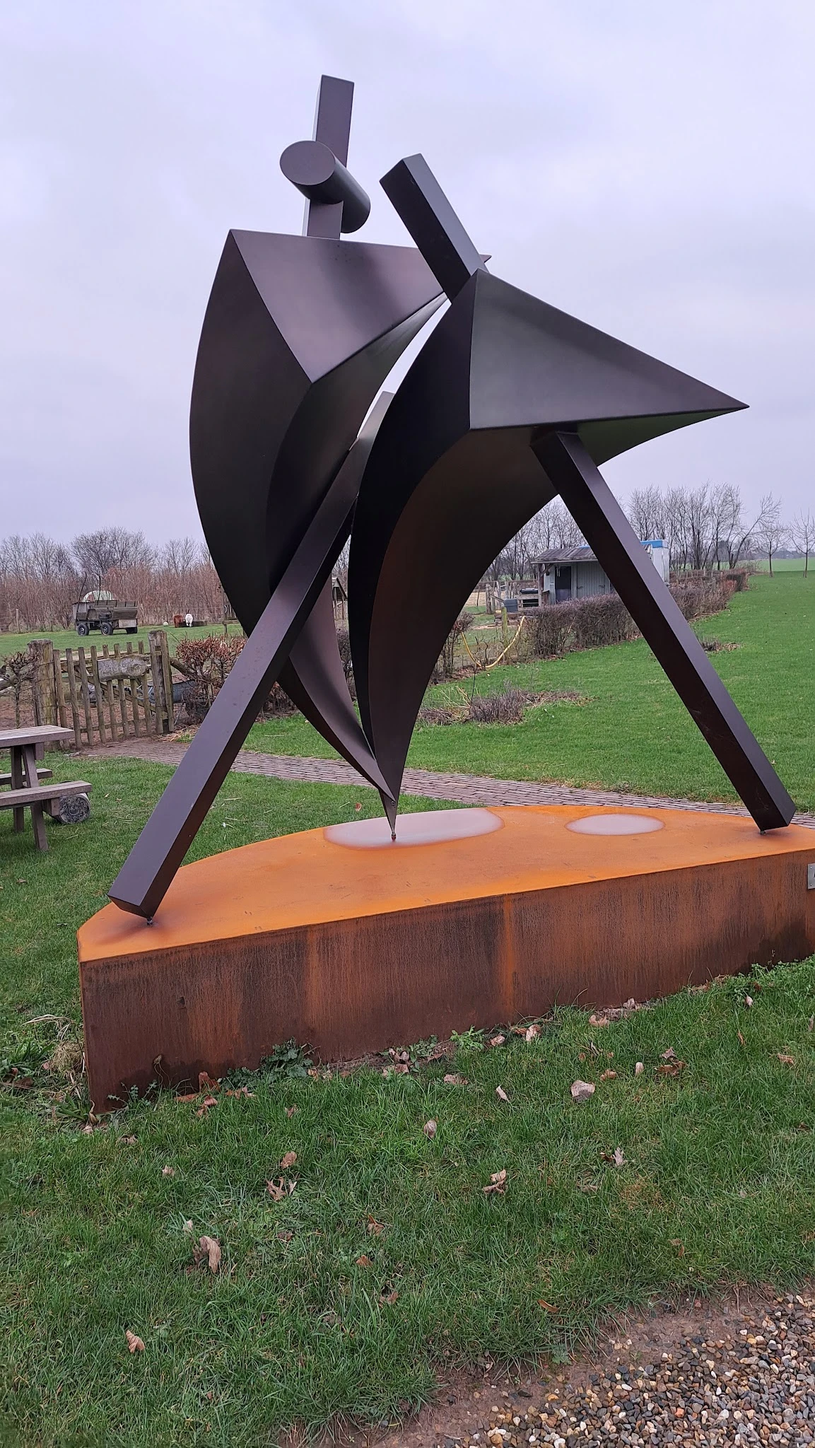 Moderne sculptuur met zwarte metalen vormen en oranje basis in een landelijk park met grasveld