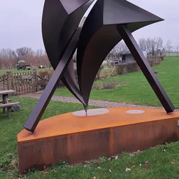 Moderne sculptuur met zwarte metalen vormen en oranje basis in een landelijk park met grasveld