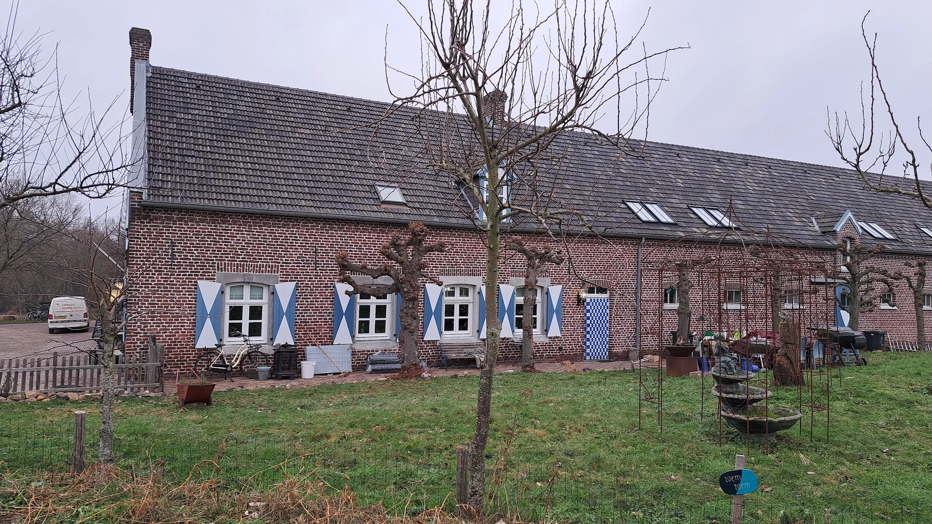 Bakstenen boerderij met blauwe luiken en witte ramen, voortuin met gras en kale boom
