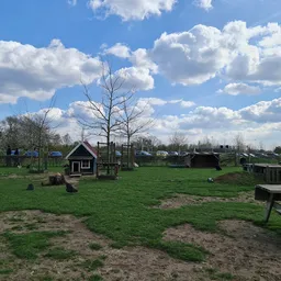 Groen terrein met speeltoestellen, houten huisjes en bankjes onder bewolkte hemel