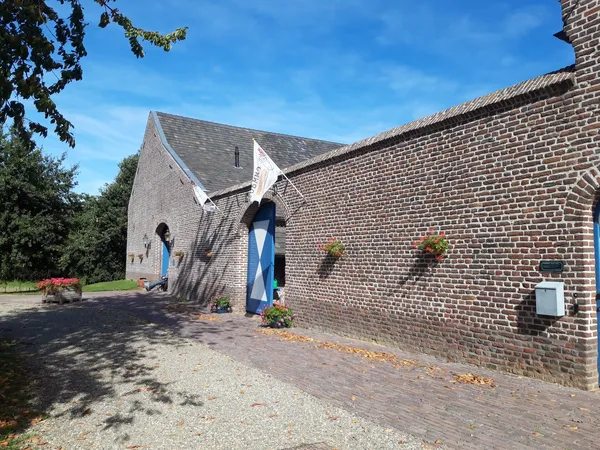 Bakstenen boerderijgebouw met blauwe deuren en ramen, omgeven door groen en straatverharding