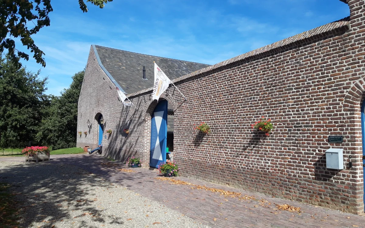 Bakstenen boerderijgebouw met blauwe deuren en ramen, omgeven door groen en straatverharding