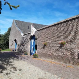 Bakstenen boerderijgebouw met blauwe deuren en ramen, omgeven door groen en straatverharding
