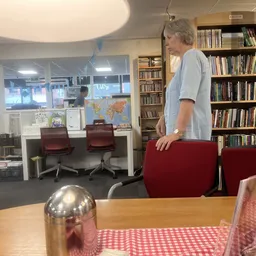 Interieur van kringloopwinkel met boekenkasten, tafel met rode stoelen en vrouw staande bij tafel