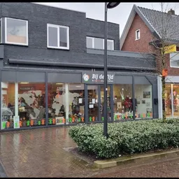 Donkerblauwe gevel van kringloopwinkel met grote etalageruiten, winkelnaam 'Bij de Tijd' zichtbaar, kleurrijke decoratie in r
