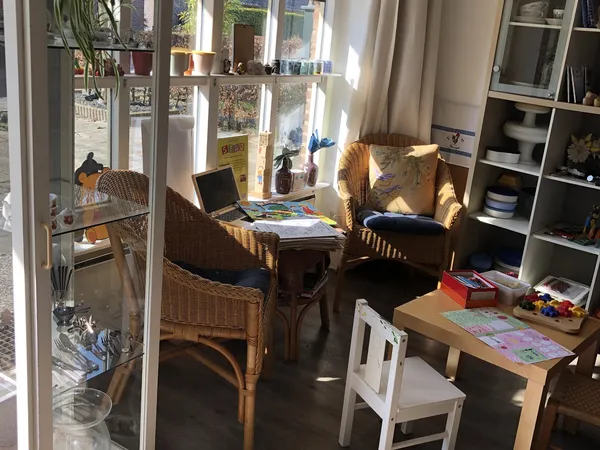 Zonnig interieur met rieten stoelen, lage tafel met laptops, boekenrek en diverse spullen langs ramen met planten