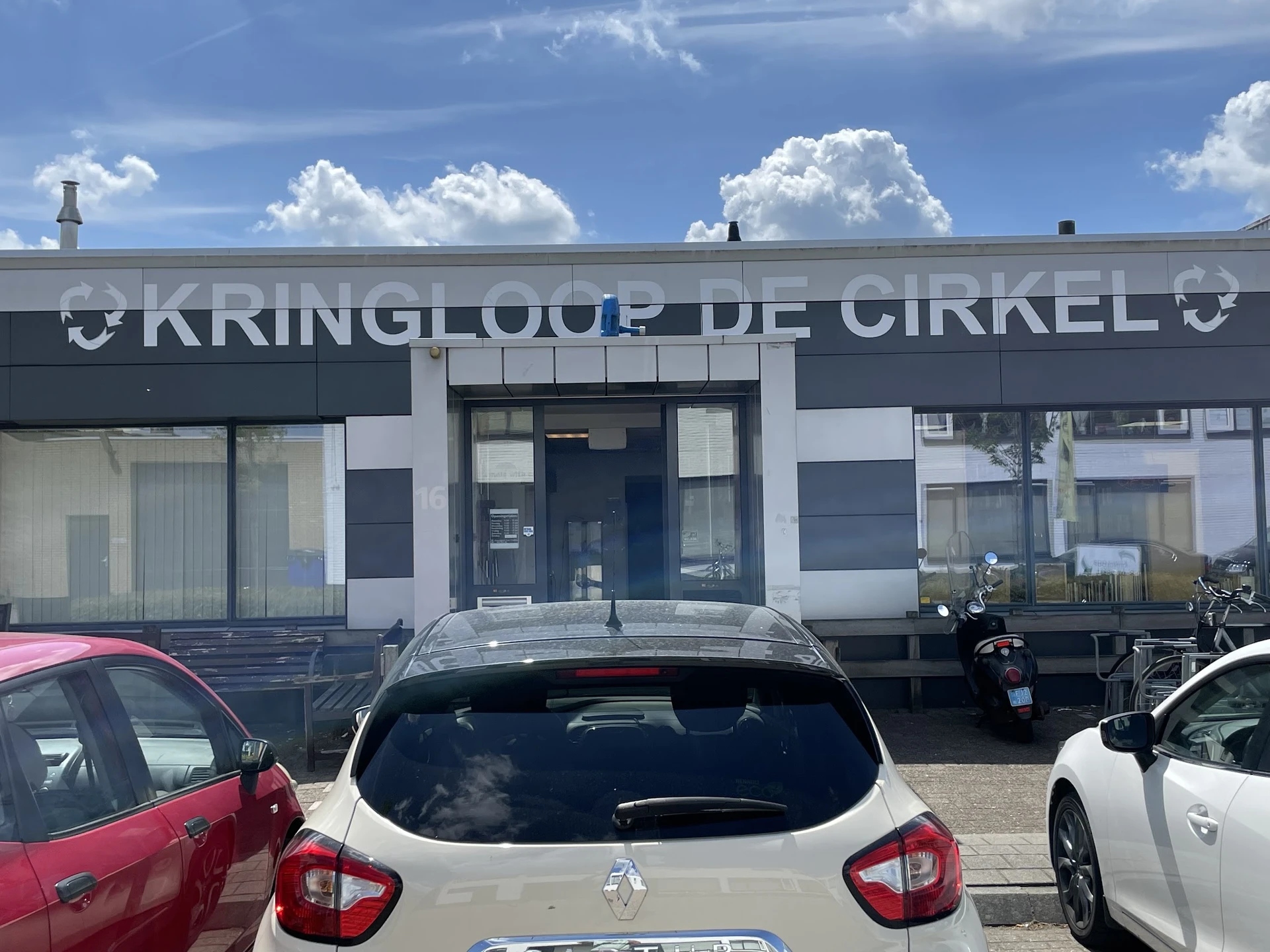 Gevel van Kringloop De Cirkel met winkelnaam op donkerblauw pand, geparkeerde auto's ervoor
