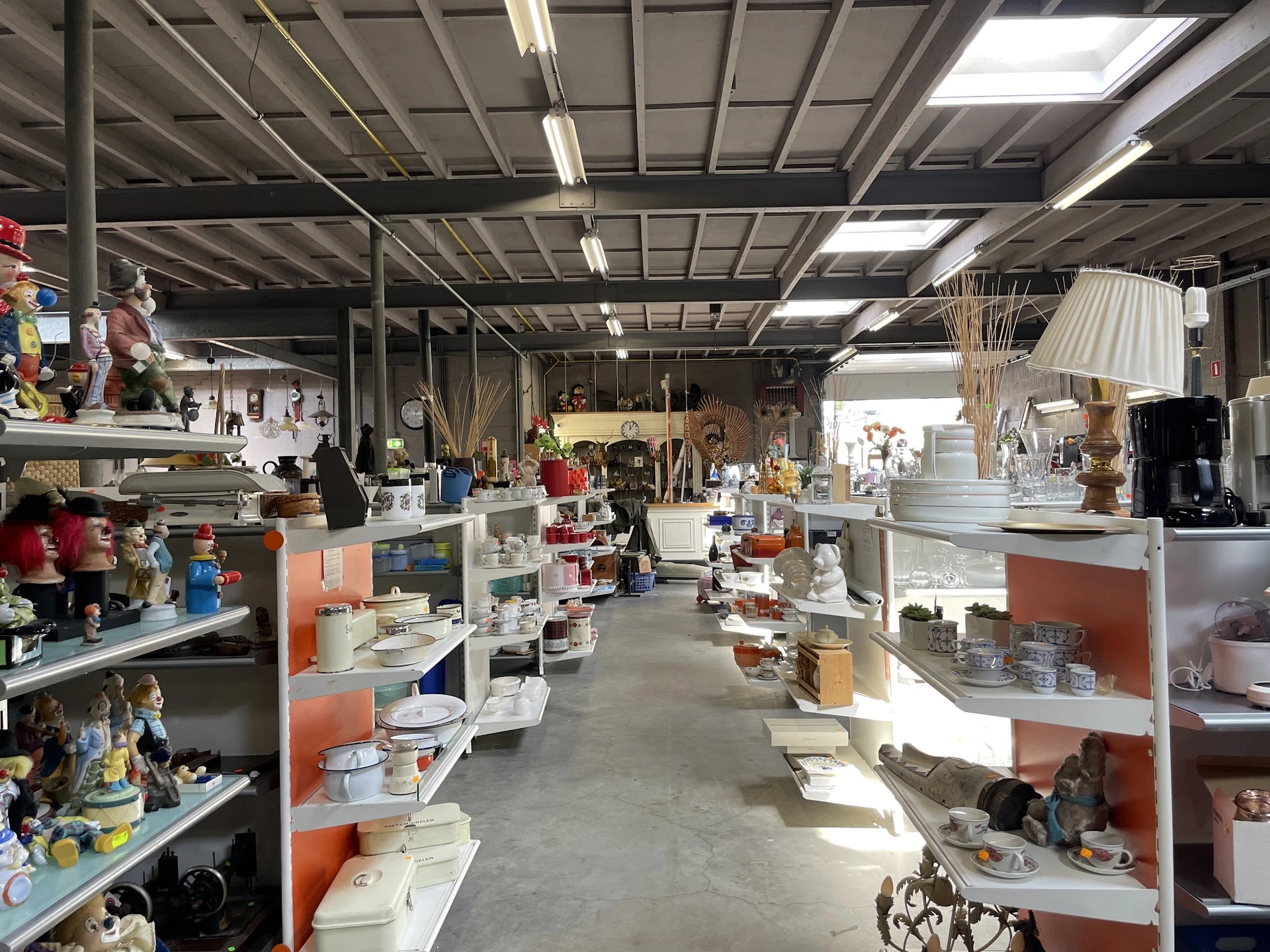 Binnenaanzicht van kringloopwinkel met stellingen vol huishoudelijke spullen, decoratie en diversen aan beide zijden van het 