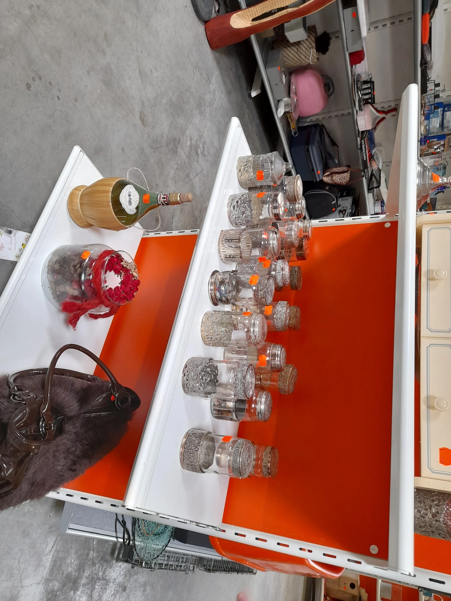 Uitgestalde items in kringloopwinkel: glazen potten met oranje dop, houten flessenhouder, rode kroon-decoratie en zwarte tas