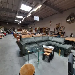Winkelvloer met grijze hoekbank, glazen salontafel en diverse meubels en decoratiestukken in het magazijn