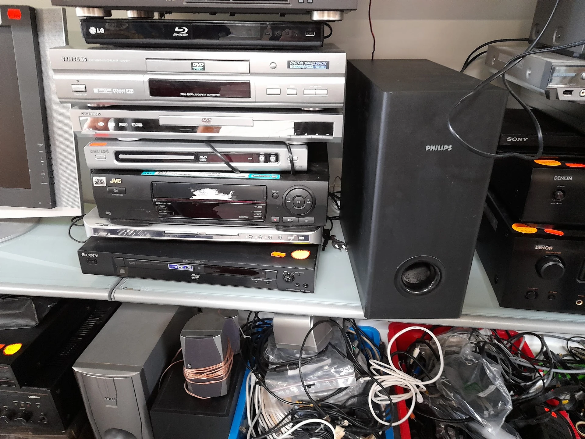 Gestapelde elektronische apparaten: blu-rayspeler, videorecorder, dvd-spelers en speakers in een kringloopwinkel