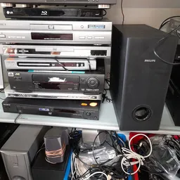 Gestapelde elektronische apparaten: blu-rayspeler, videorecorder, dvd-spelers en speakers in een kringloopwinkel