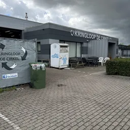 Grijze voorgevel van Kringloop De Cirkel met logo, ingang en terras met stoelen