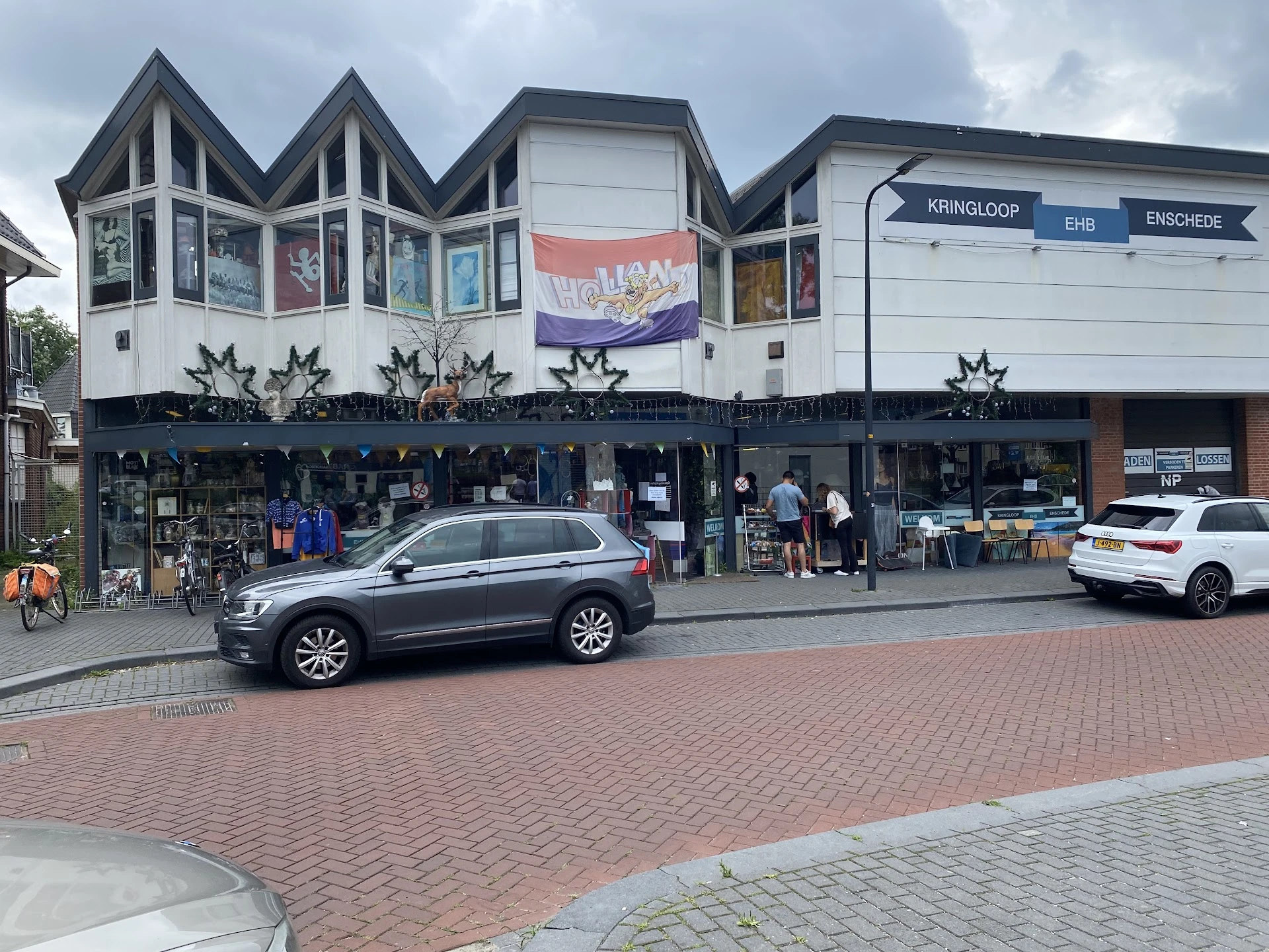 Voorgevel van kringloopwinkel met witte en zwarte gevel, grote ramen, winkelnaam bovenaan zichtbaar, geparkeerde auto's op st