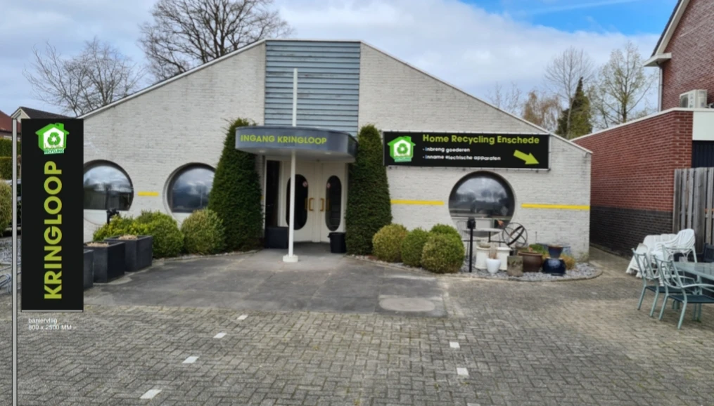 Witgeschilderde gevel van kringloopwinkel met twee deuren, ronde ramen en groene beplanting
