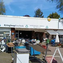 Witte gevel van kringloopwinkel met naam en logo, omgeven door uitgestalde tweedehands spullen op het terrein