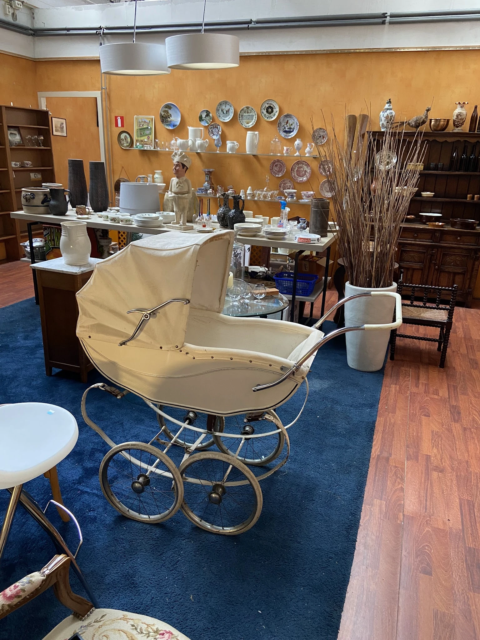 Vintage kinderwagen met crèmekleurig canvas en zilveren frame op blauwe vloer in kringloopwinkel