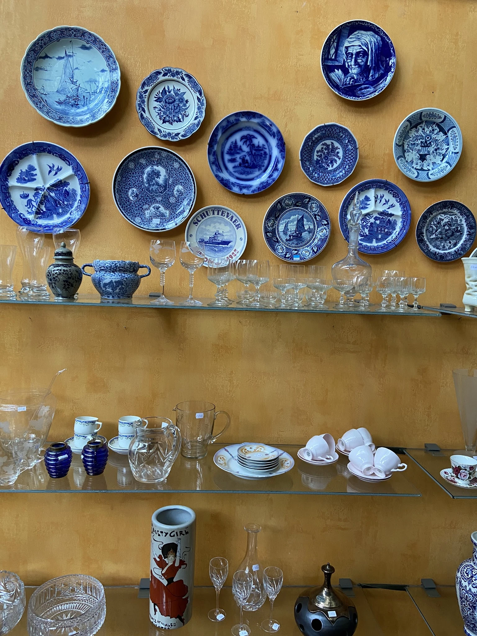 Glazen en servies uitgestald op twee houten schappen, met blauwe borden aan de muur