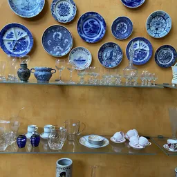 Glazen en servies uitgestald op twee houten schappen, met blauwe borden aan de muur