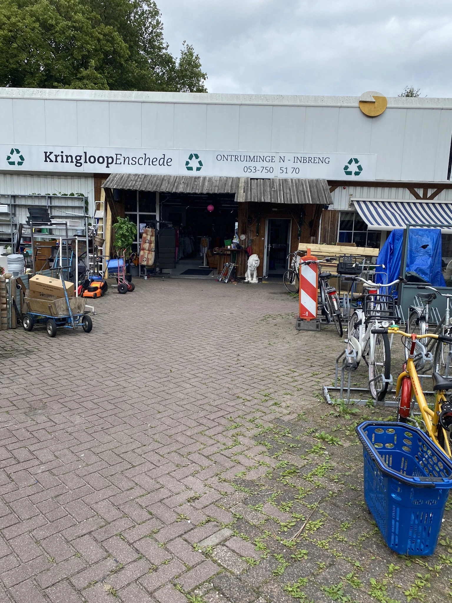 Voorgevel van kringloopwinkel met ingang, houten luifel en fietsen ernaast