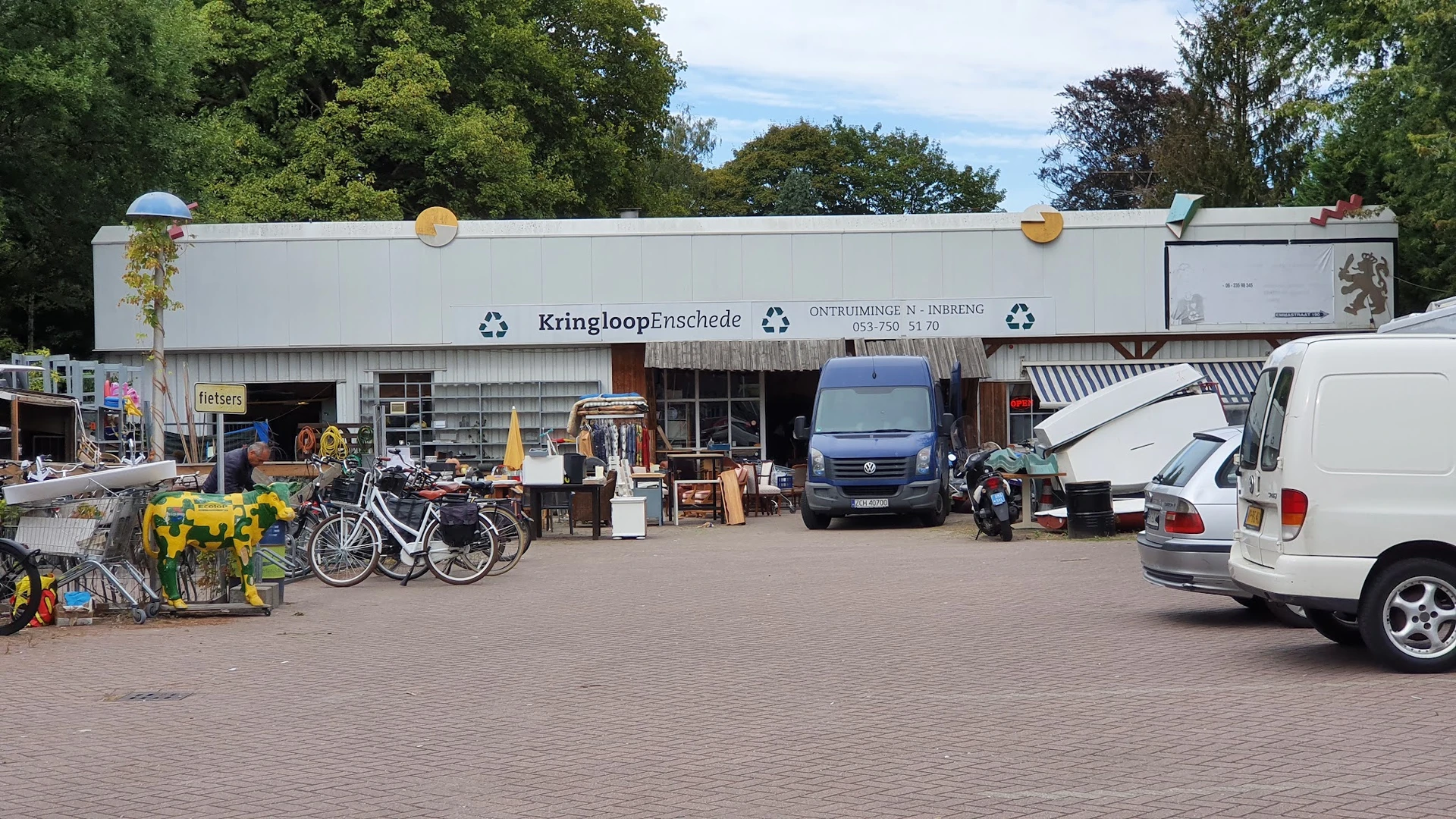 Voorgevel van Kringloop Enschede met winkelnaam op grijze gevelbekleding, parkeerplaats met fietsen en voertuigen.