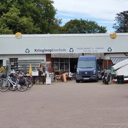 Voorgevel van Kringloop Enschede met winkelnaam op grijze gevelbekleding, parkeerplaats met fietsen en voertuigen.