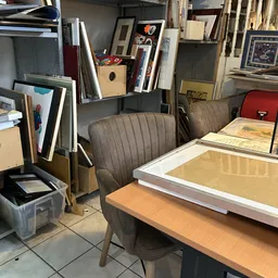 Binnenaanzicht van kringloopwinkel met bureaus, stoelen en ingelijste kunstwerken tegen muren