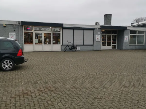 Gevel van kringloopwinkel met witte en grijze pui, naam zichtbaar op raam, parkeerplaats met auto en fiets ervoor