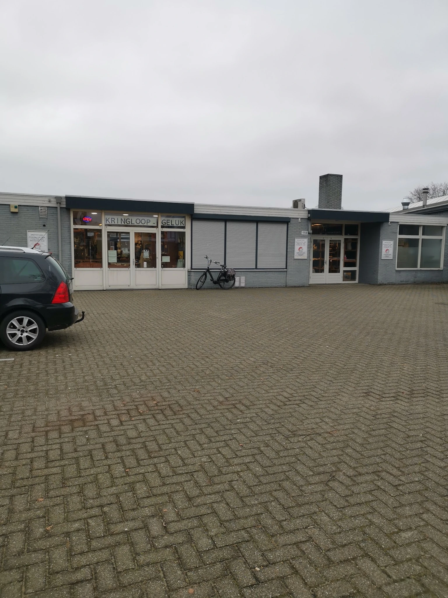 Gevel van kringloopwinkel met witte en grijze pui, naam zichtbaar op raam, parkeerplaats met auto en fiets ervoor