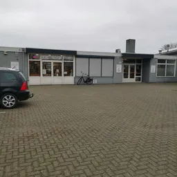 Gevel van kringloopwinkel met witte en grijze pui, naam zichtbaar op raam, parkeerplaats met auto en fiets ervoor