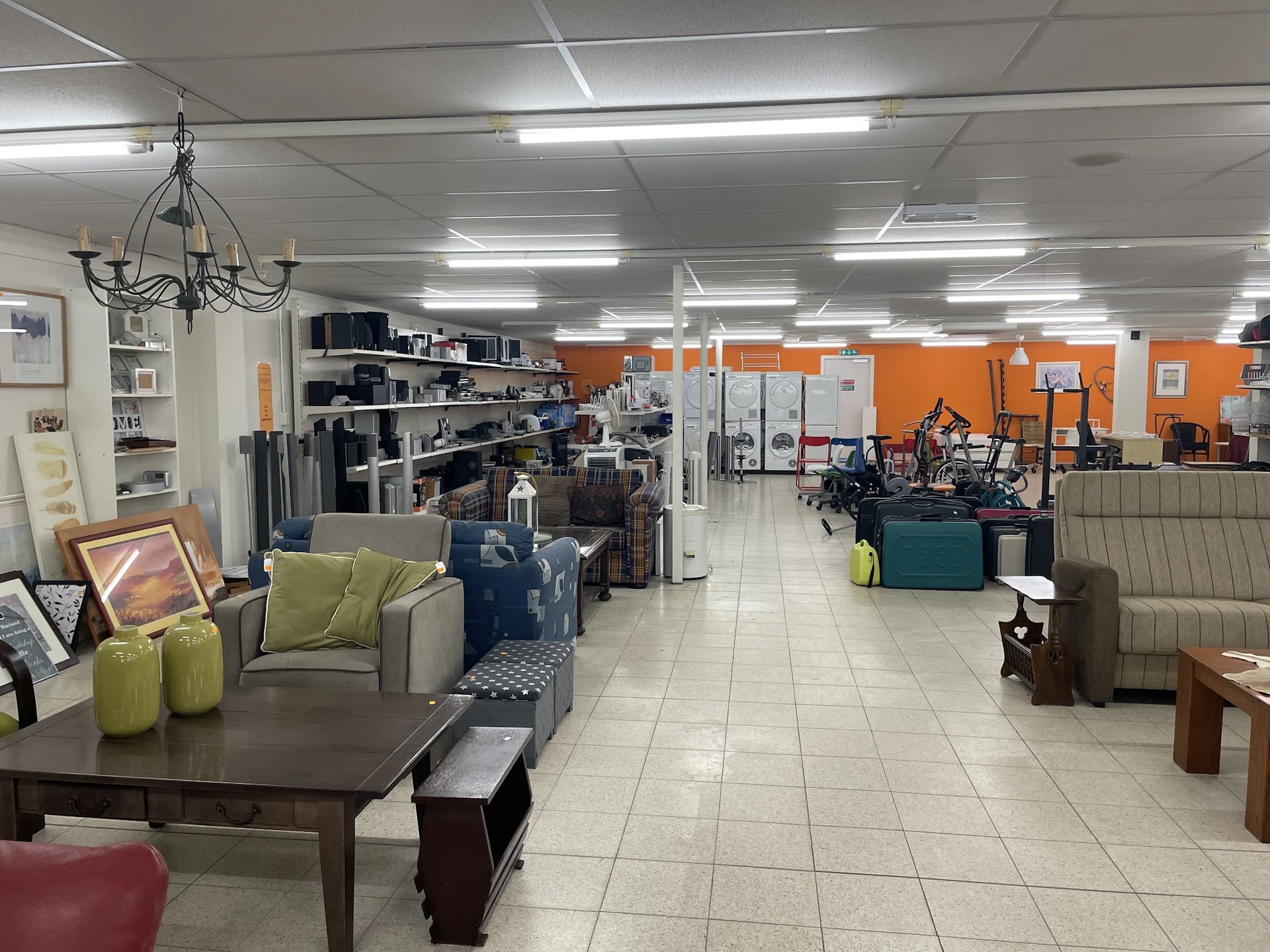 Ruime kringloopwinkel met meubels, huishoudspullen en decoratie uitgestald, oranje muren en fluorescente verlichting