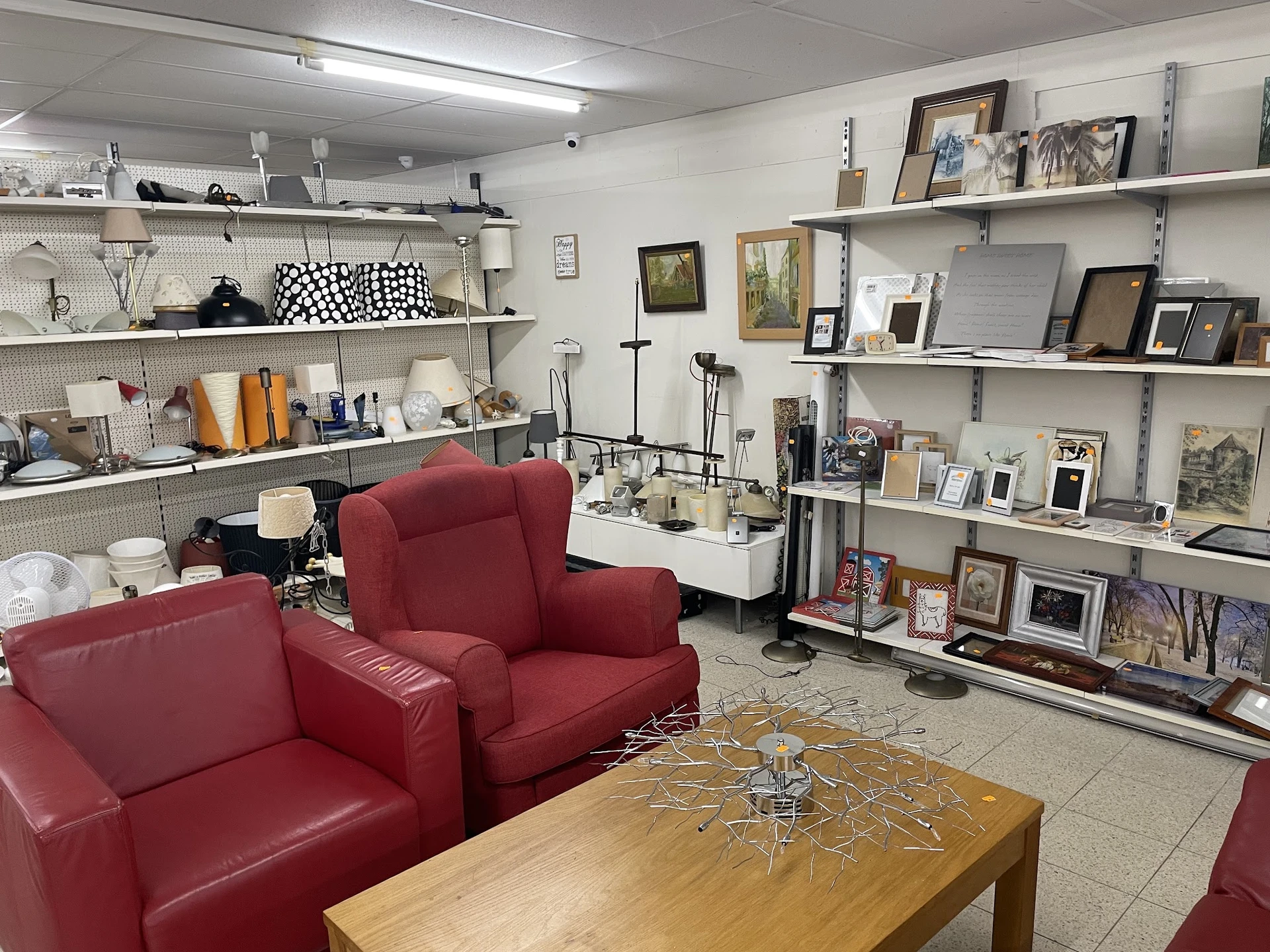 Winkelvloer met rode fauteuils, houten tafel, en witte stellingen vol met huishoudspullen, foto's en decoratie