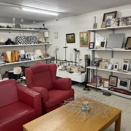 Winkelvloer met rode fauteuils, houten tafel, en witte stellingen vol met huishoudspullen, foto's en decoratie