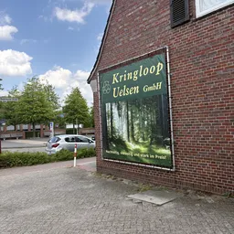 Groen winkelbordje van Kringloop Uelsen GmbH aan bakstenen gevel met parkeerplaats en auto's op achtergrond
