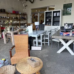Winkelinterieur met witte eetkamertafel en stoelen, rieten stoel op voorgrond, stellingen met huishoudartikelen en decoratie 